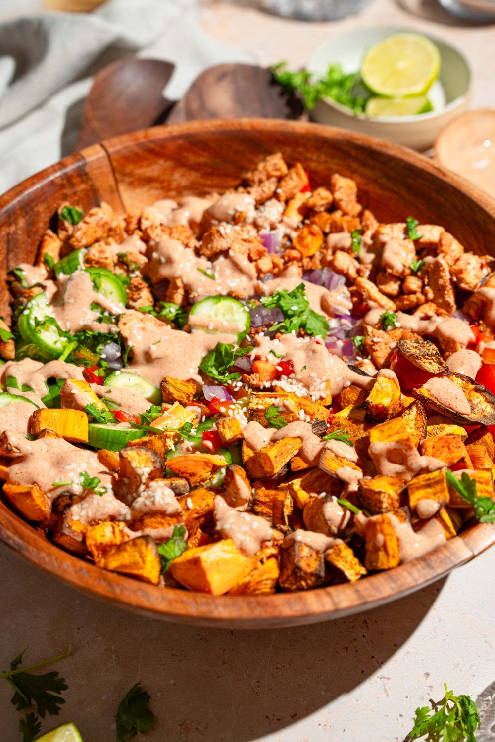 A large wooden bowl with hot honey chicken bowl including sweet potato, chicken, cucumber, onion, and bell peppers drizzled with hot honey sauce. The bowl is on a tan counter with small bowls of ingredients.