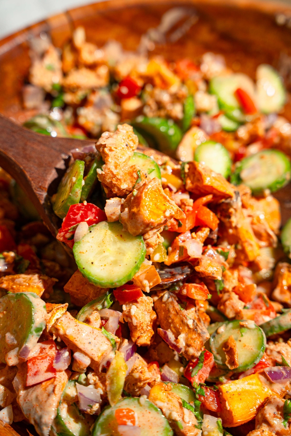 A large wooden bowl with hot honey chicken bowl including sweet potato, chicken, cucumbers, onion, and bell pepper tossed in a hot honey sauce. A salad spoon is scooping the mixture.