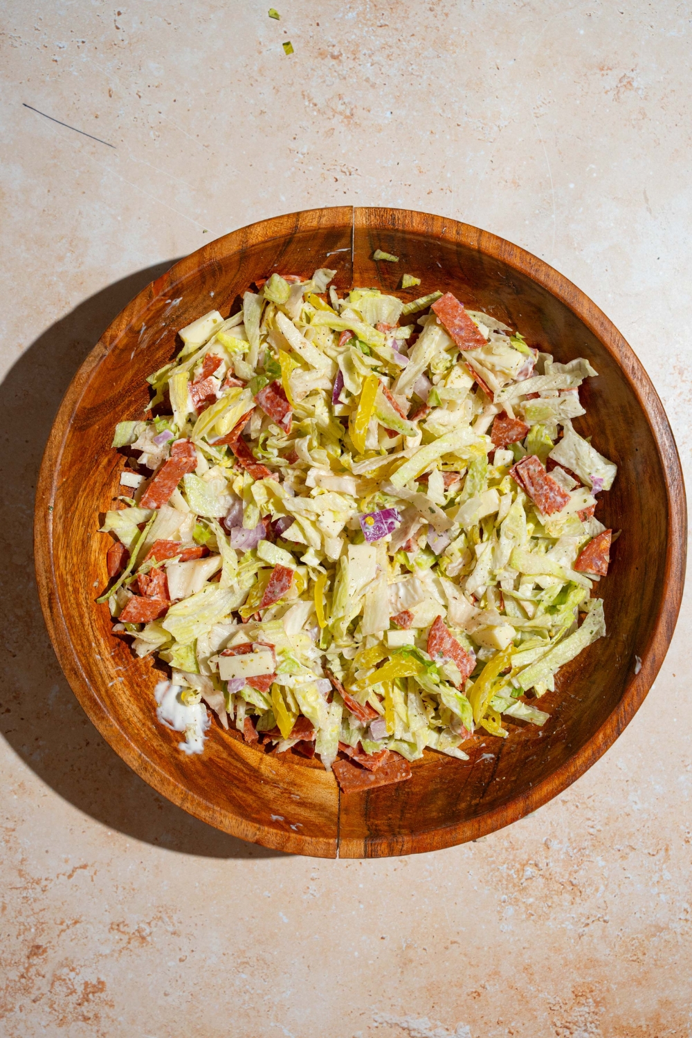 A large wooden bowl with chopped lettuce, salami, pepperoni, banana peppers, red onion, provolone cheese, and more. The bowl is on a tan counter.