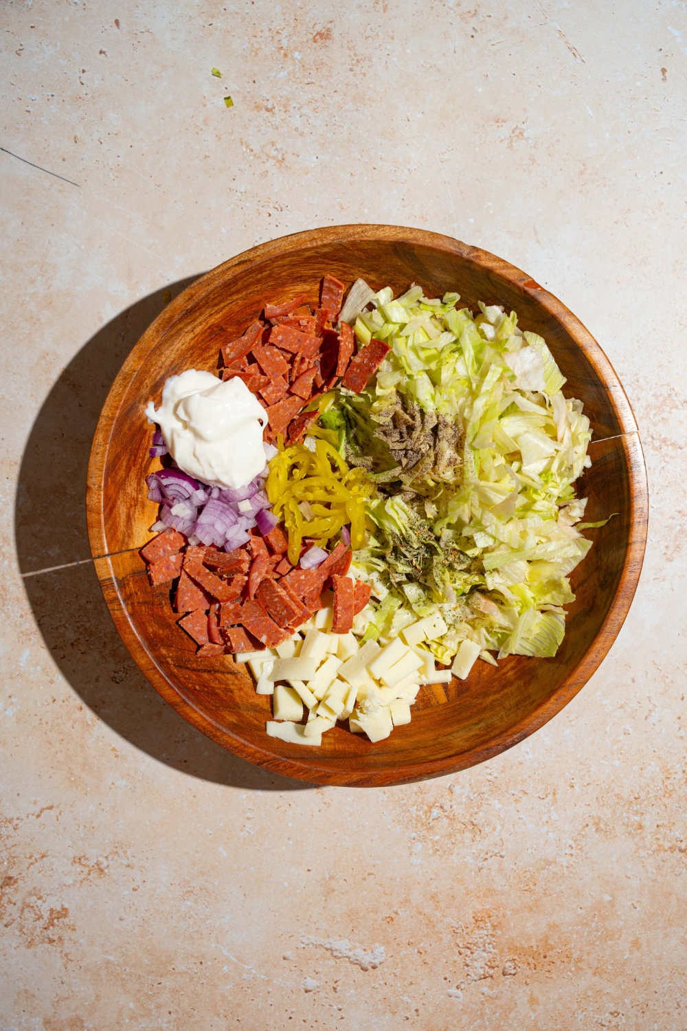 A large wooden bowl with ingredients to make Italian grinder dip including lettuce, salami, pepperoni, red onion, provolone cheese, mayo, and seasonings. The bowl is on a tan counter.