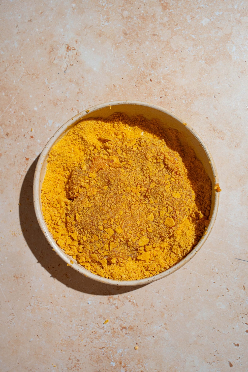 A large bowl of crushed cornflakes with a chicken patty coated in the cornflakes. The bowl is on a tan counter.