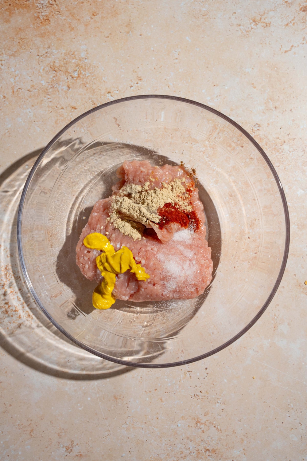 A glass bowl with ground chicken,, mustard and seasonings. The bowl is on a tan counter.