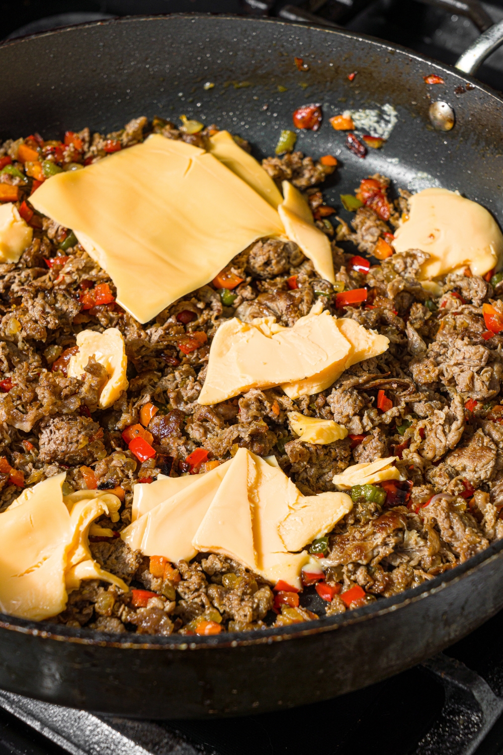 A skillet with a ground beef and pepper mixture topped with sliced cheese. The skillet is on a stove.