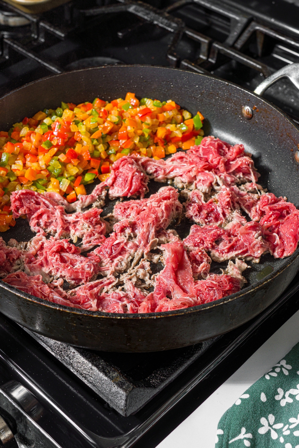 A skillet with diced peppers and uncooked ground beef. The skillet is on a stove.