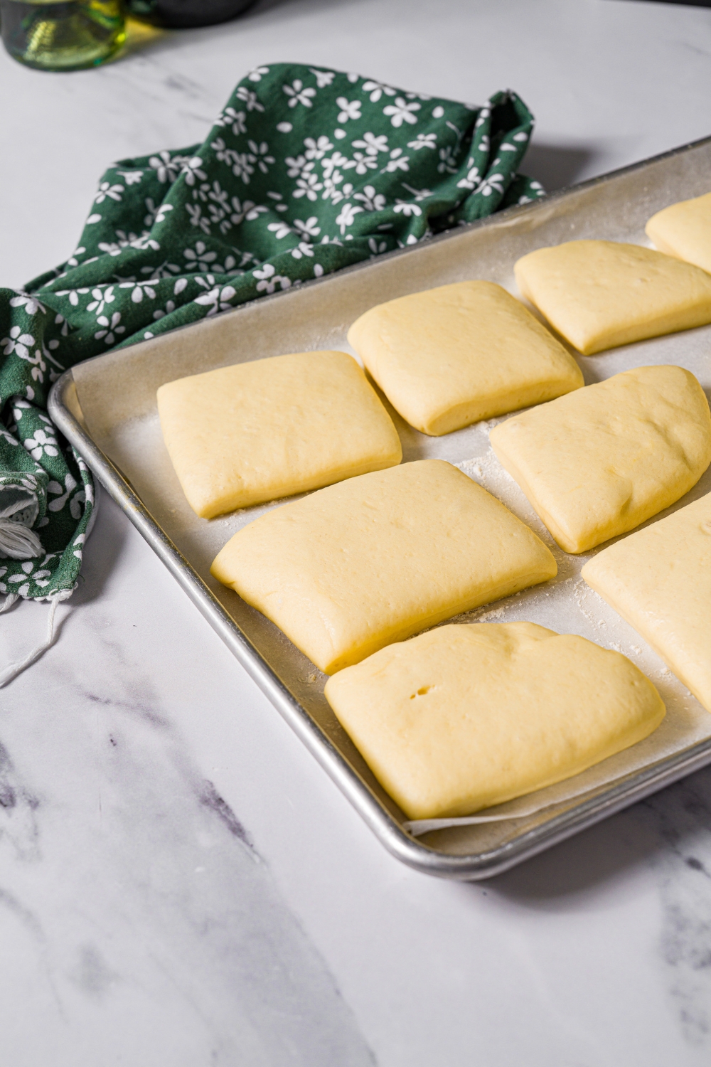 A baking sheet lined with parchment paper with slices of beignet dough proofing on the sheet. The sheet is on marble counter with a green floral napkin.