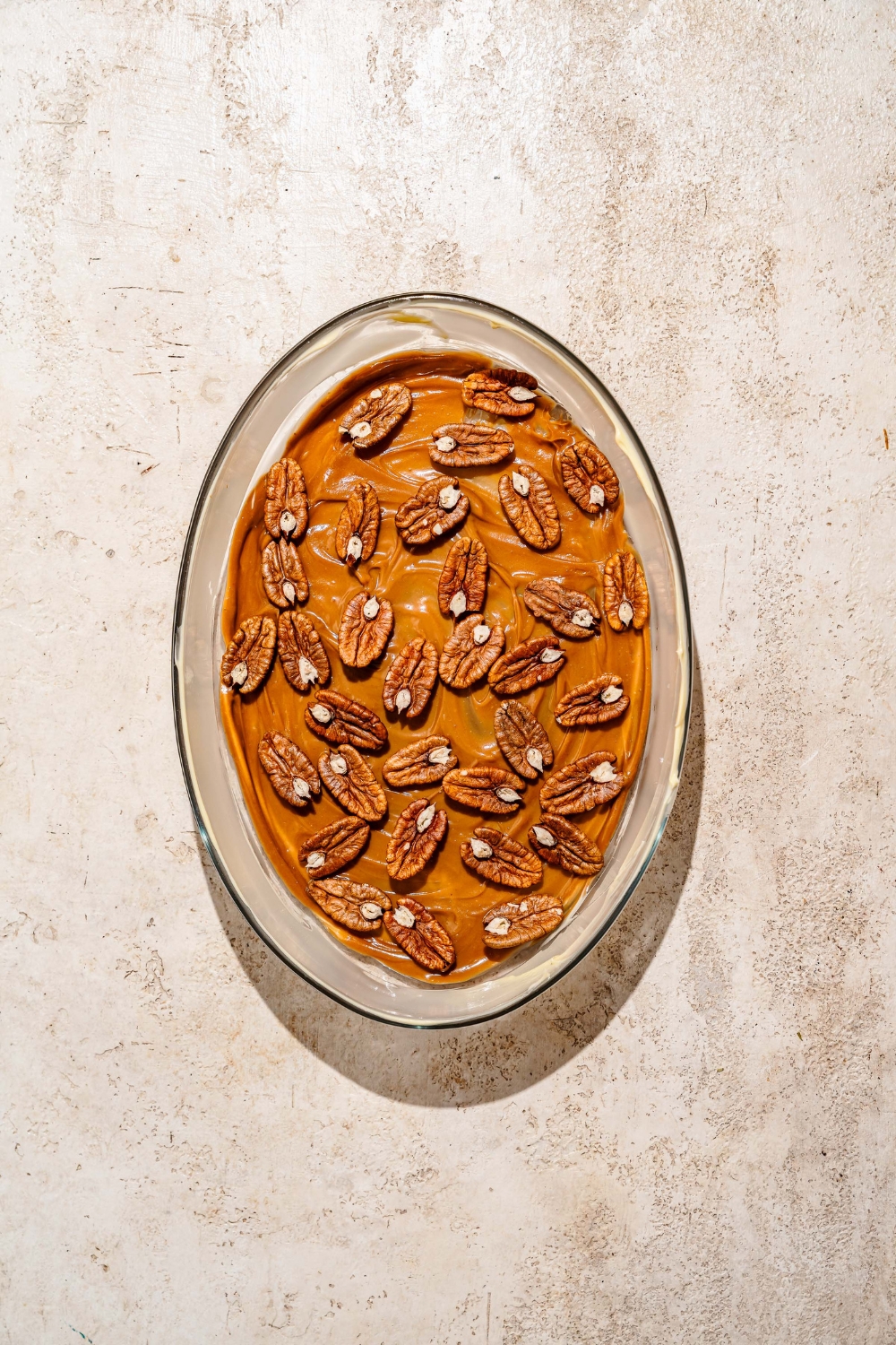 An oval baking dish with caramel mixture and pecans spread over the base. The dish is on a tan counter.