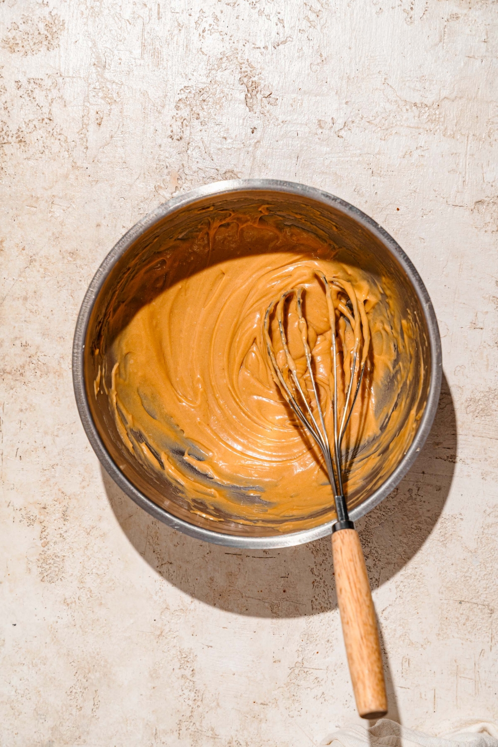 A large bowl with a whisk mixing Biscoff cookie butter cream cheese frosting. The bowl is on a tan counter.