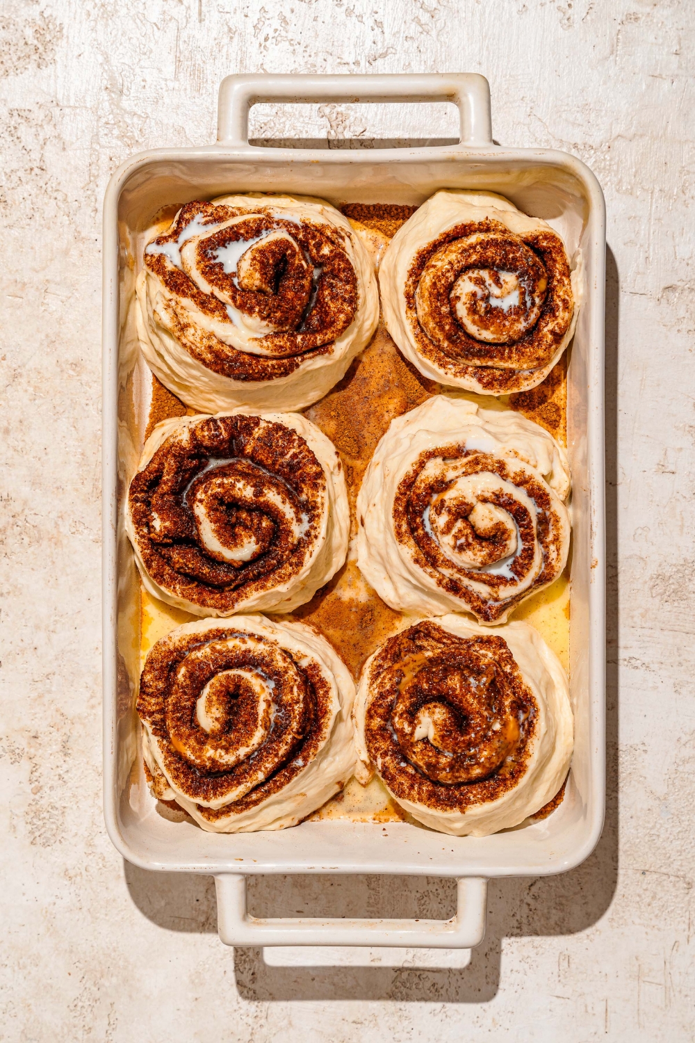 A baking pan coated with a cinnamon mixture with unbaked rolled Biscoff cinnamon rolls coated with milk and butter. The pan is on a tan counter.