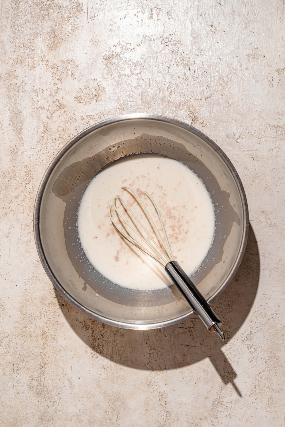 A large bowl with a whisk mixing milk, sugar, and yeast. The bowl is on a tan counter.