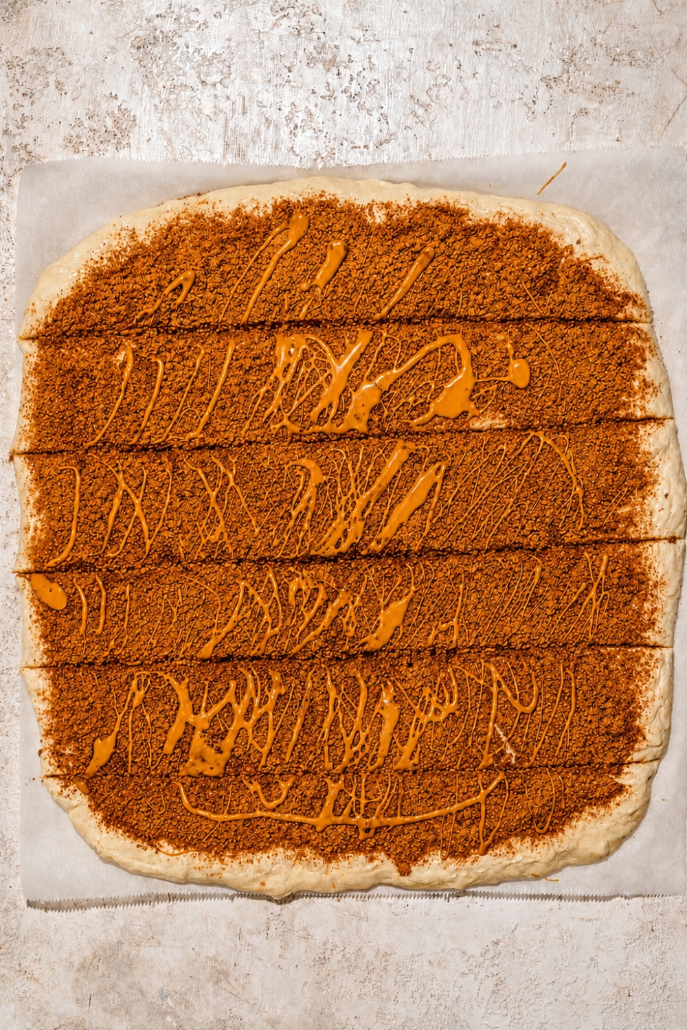 A piece of parchment paper with cinnamon roll dough rolled out into a square shape topped with cinnamon and Biscoff cookie filling and drizzled with melted Biscoff cookie butter. The dough has been sliced in to strips.