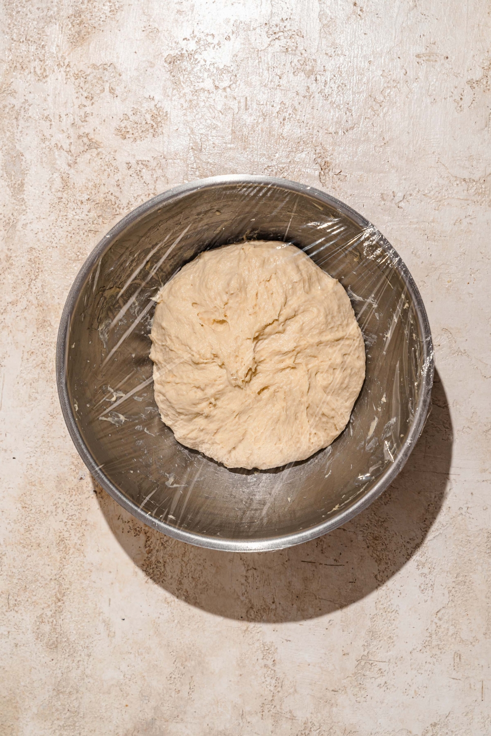 A large ball of dough in a bowl covered with plastic wrap. The bowl is on a tan counter.