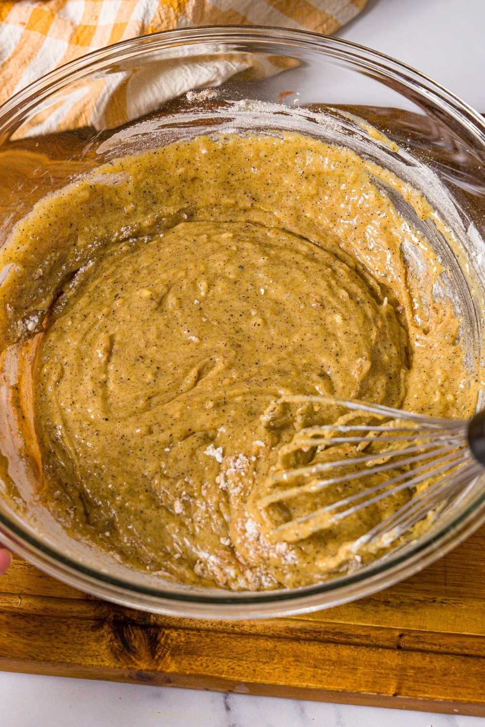 A large bowl with a whisk mixing carrot cake batter. The bowl is on a wooden board.