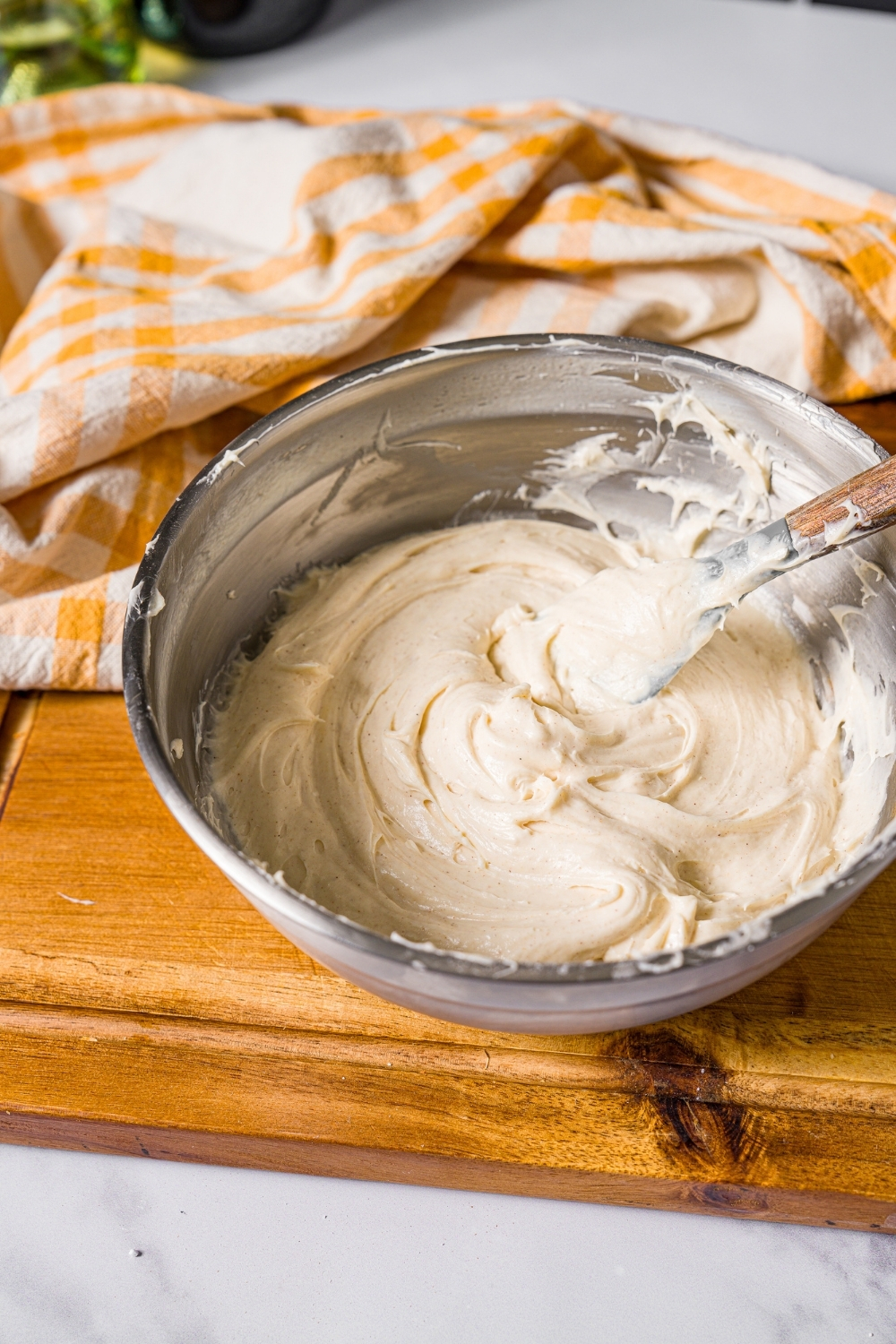 A large mixing bowl with a spatula mixing cream cheese frosting. The bowl is on a wooden board with a yellow striped napkin.