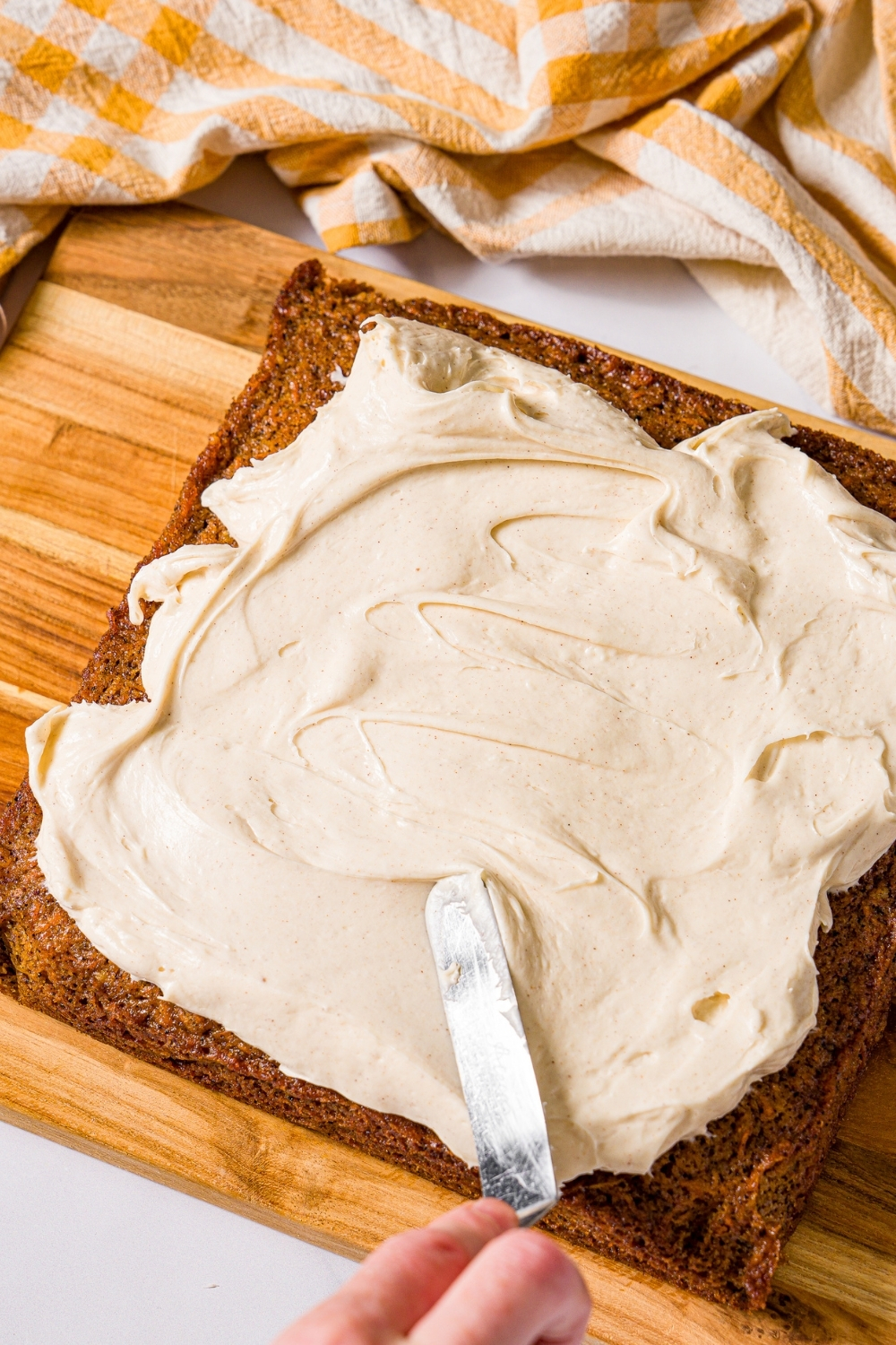 A whole brown butter carrot cake with a frosting knife spreading cream cheese frosting over the cake. The cake is on a wooden board with a yellow striped napkin.