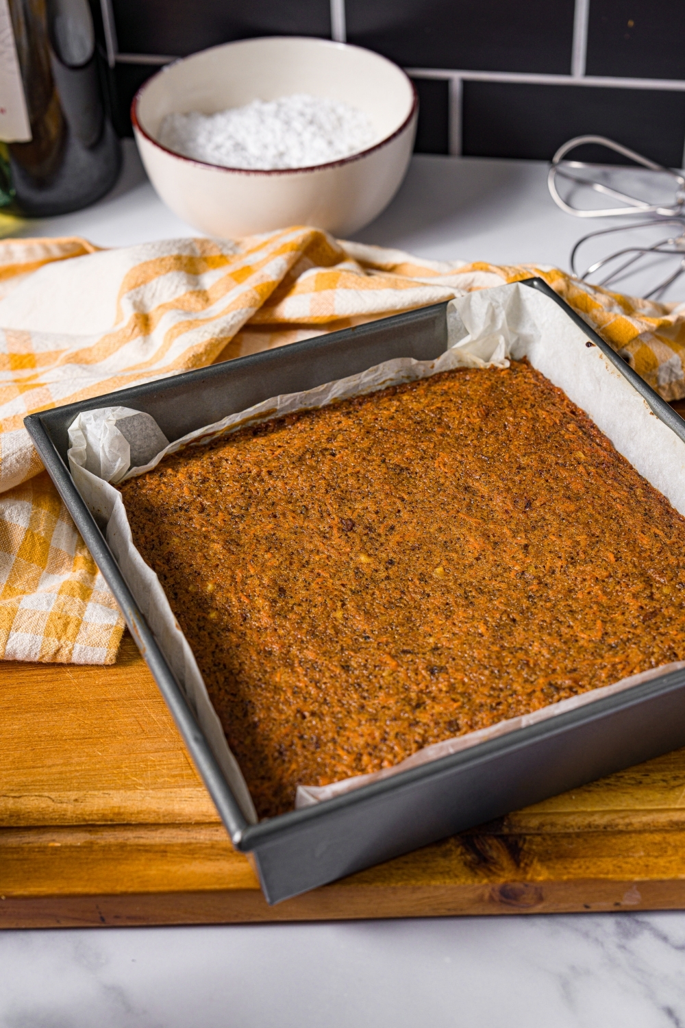 A baking pan lined with parchment paper with baked brown butter carrot cake. The pan is on a wooden board with a yellow striped napkin.