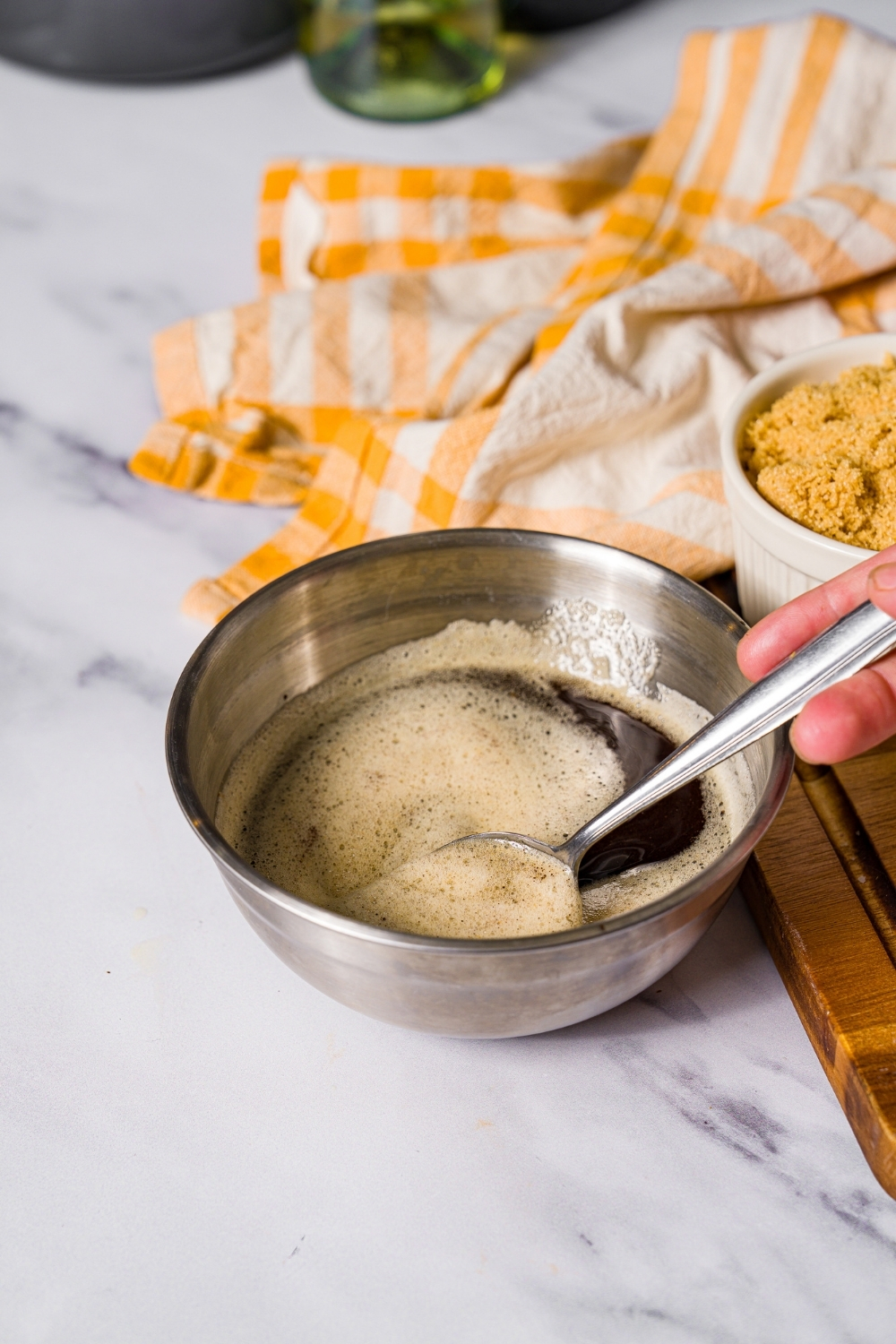 A small bowl with a spoon stirring brown butter. The bowl is on a marble counter with a yellow striped napkin.