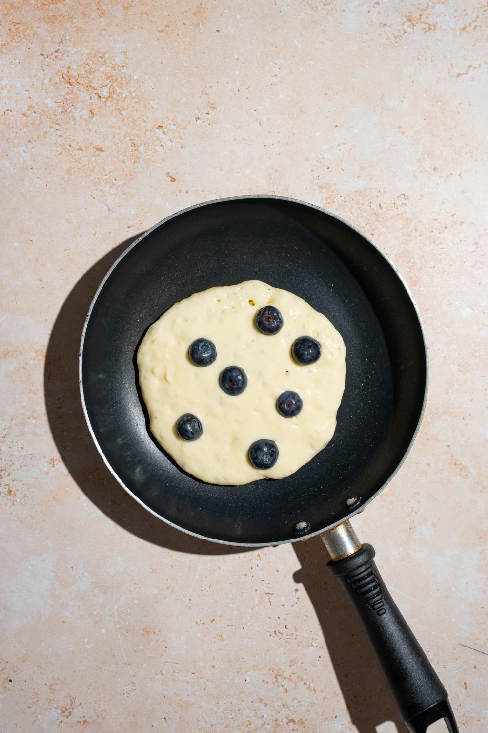 A skillet with an unflipped blueberry pancake. The skillet is on a tan counter.