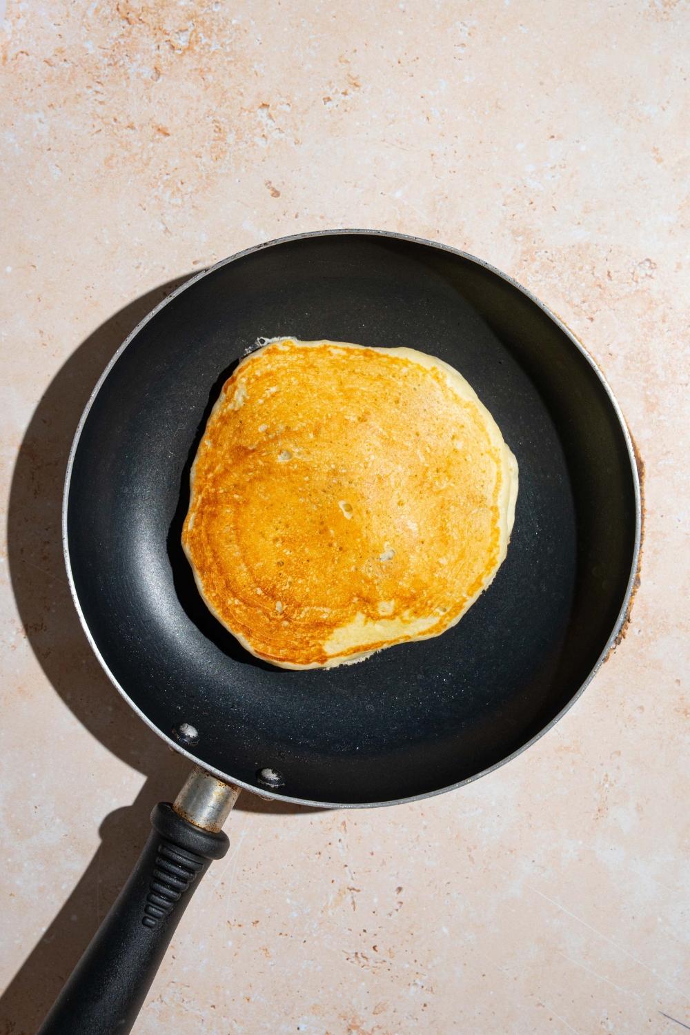 A skillet with a flipped blueberry pancake. The skillet is on a tan counter.