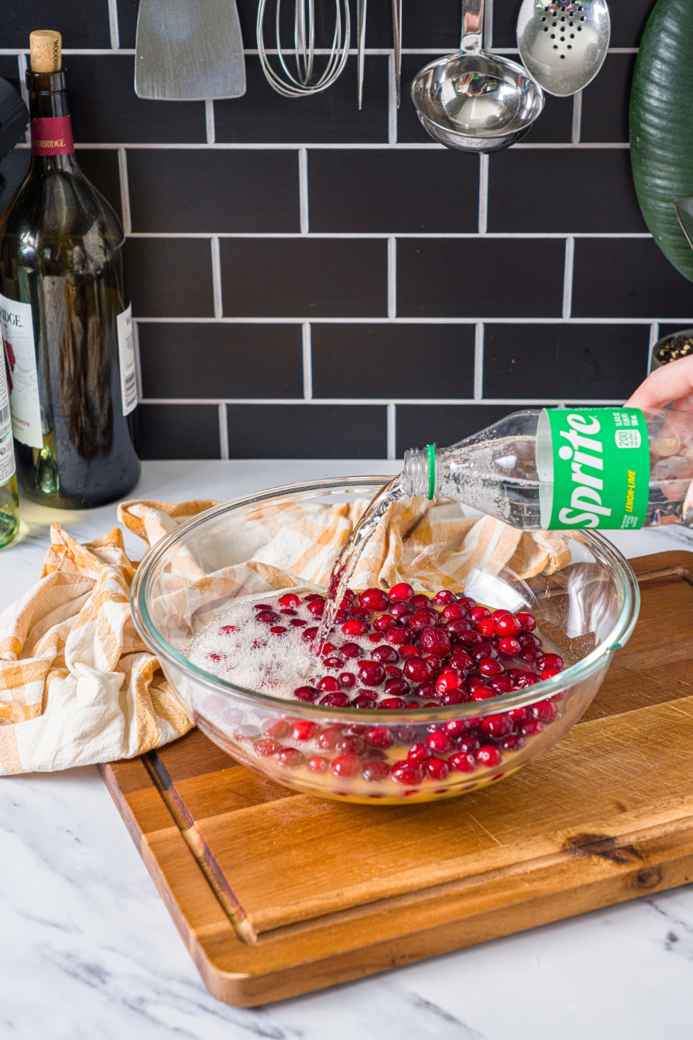 A large glass bowl with sprite being added to a cranberry mixture. The bowl is on a wooden board with a yellow striped napkin.