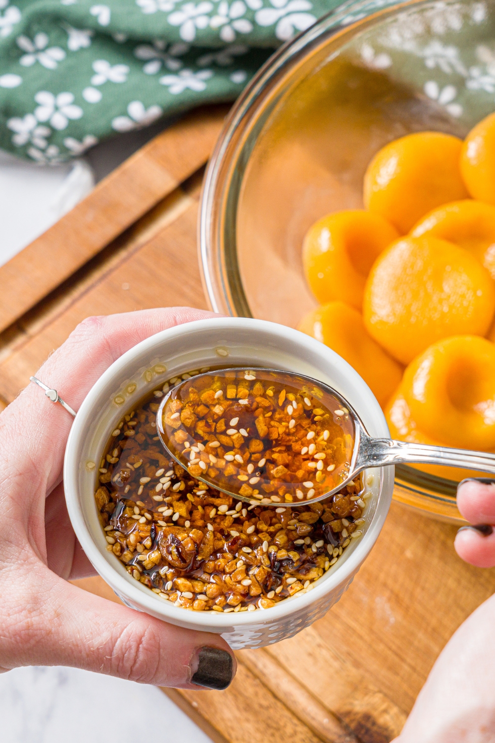 A small ramekin of chili crisp sauce with a spoon stirring the mixture. There is a bowl of carrot button noodles blurred in the background.
