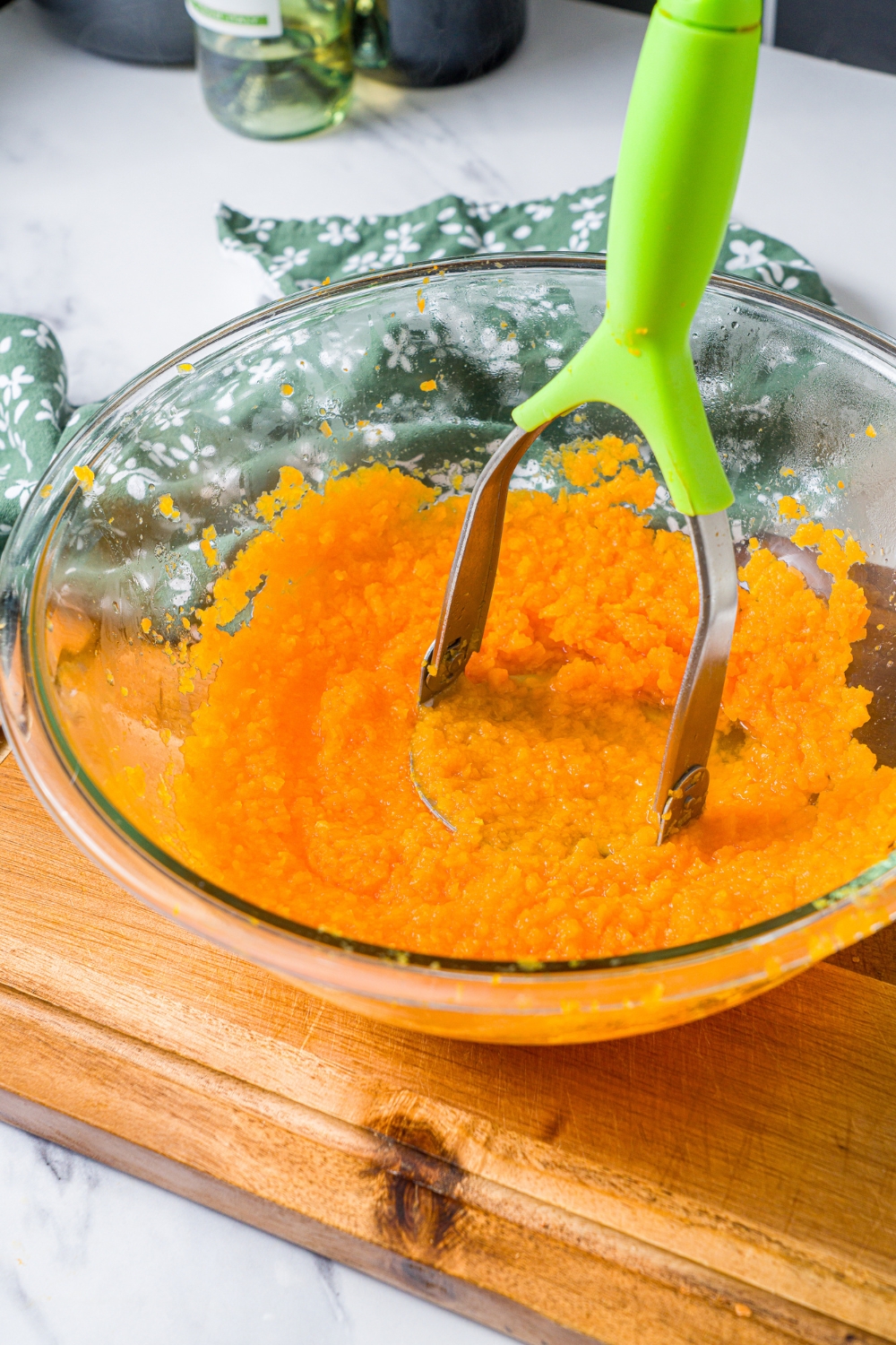 A glass bowl with a masher mashing boiled carrots. The bowl is on a wooden board on a marble counter with a green floral napkin.