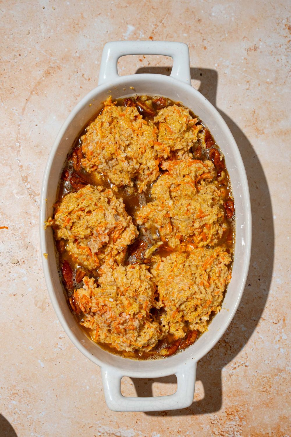 An oval dish with a pecan pie filling base topped with carrot cake cobbler dough on top. The dish is on a tan counter.