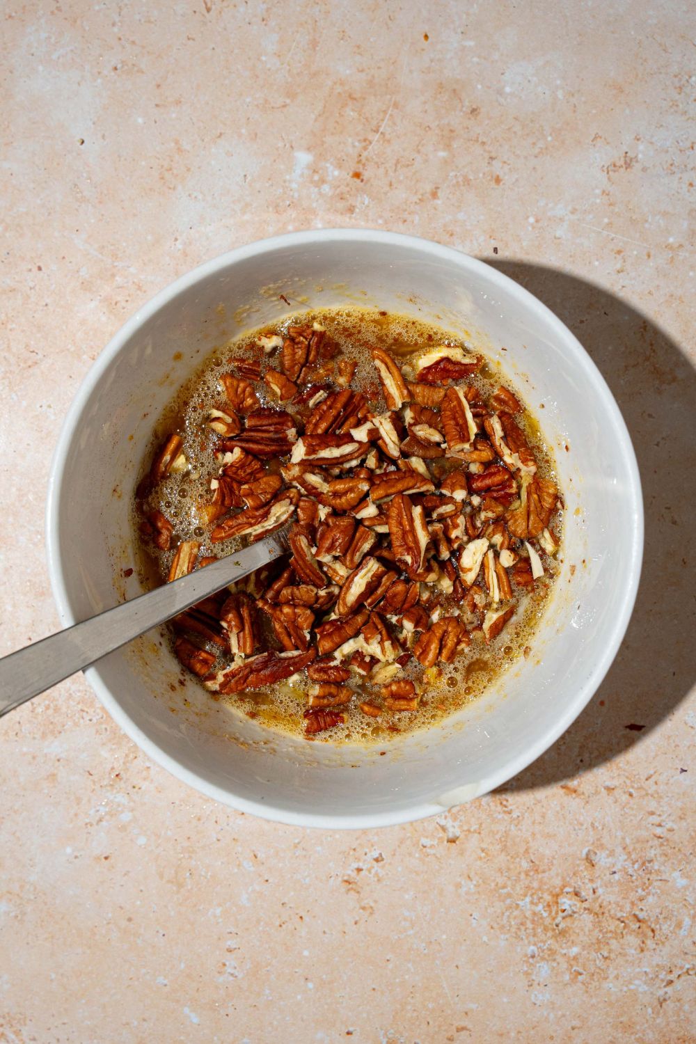 A large bowl with a spoon mixing ingredients to make pecan pie filling including eggs, corn syrup, brown sugar, cinnamon, pecans, and more. The bowl is on a tan counter.