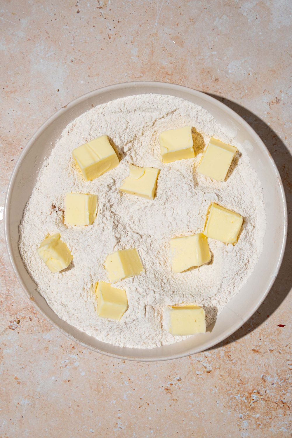 A large bowl of dry ingredient mixture topped with cubed cold butter. The bowl is on a tan counter.