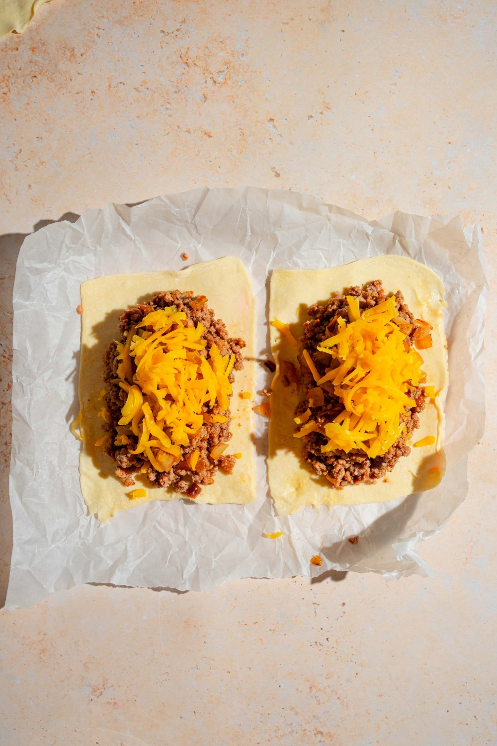 A baking sheet lined with parchment paper with two open puff pastry shells topped with ground beef, onions, and cheese. The sheet is on a tan counter.