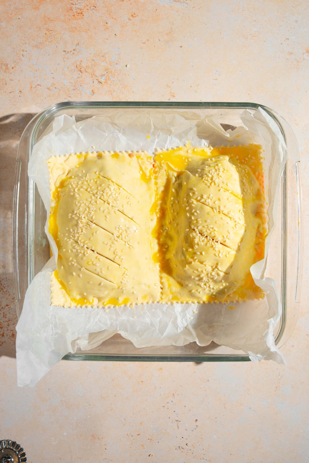 A glass baking dish lined with parchment paper with two sealed unbaked cheeseburger hot pockets. The dish is on a tan counter.