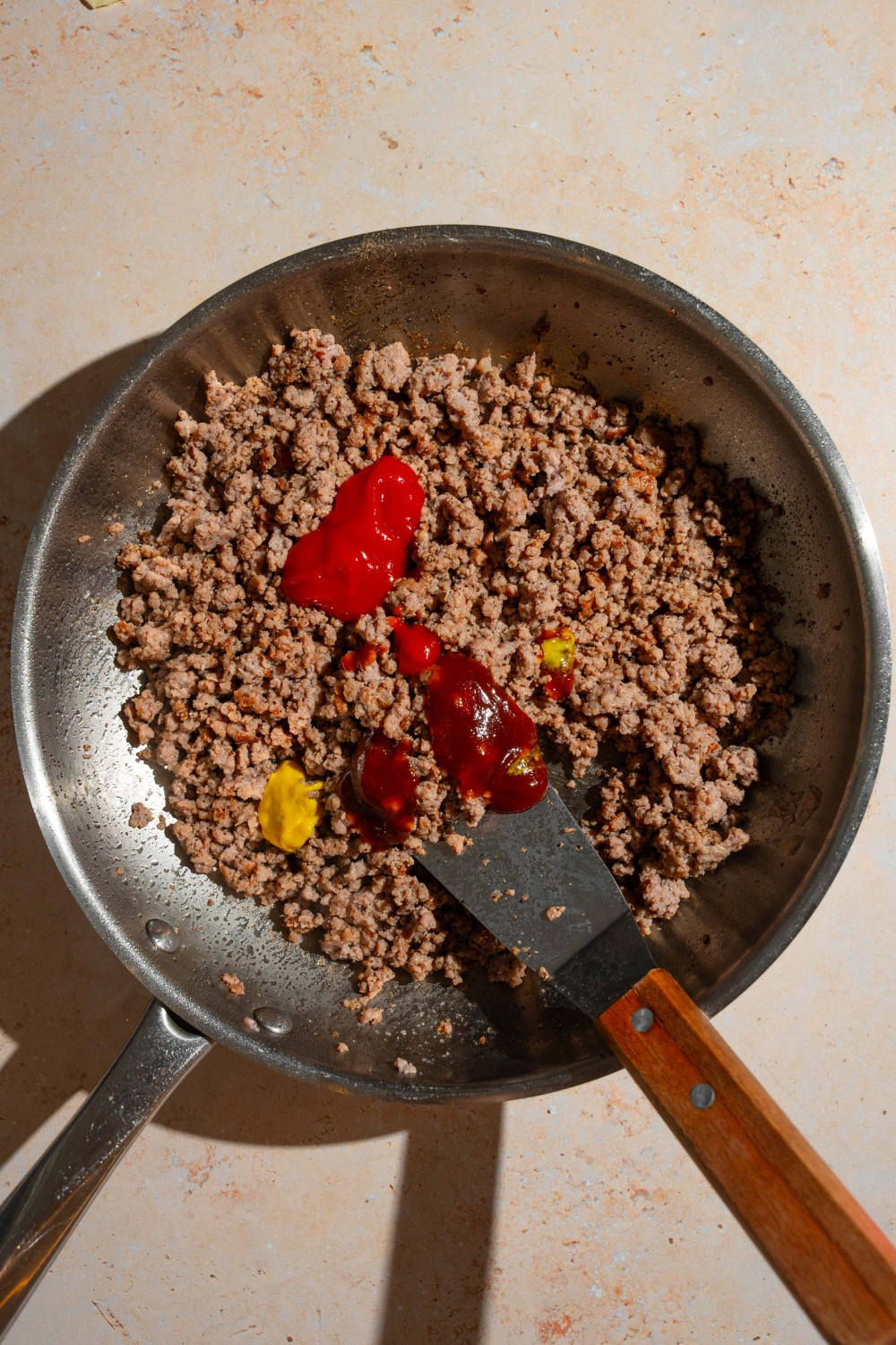 A skillet with a spatula mixing sauces and seasonings into ground beef. The skillet is on a tan counter.