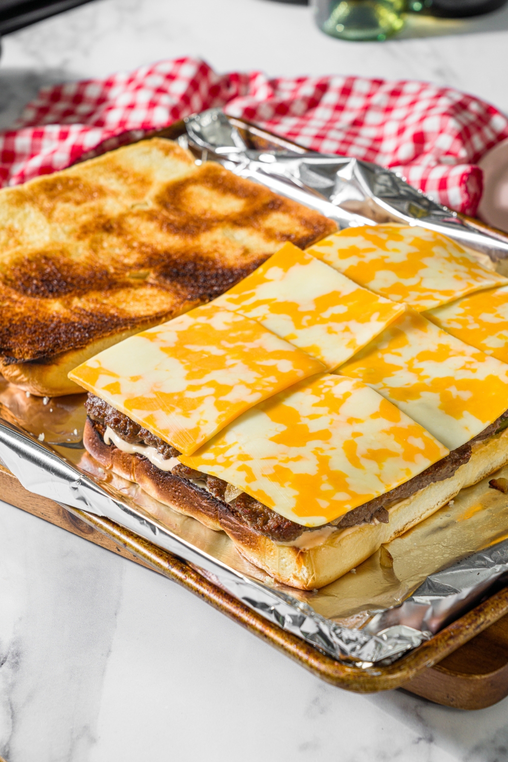 A baking sheet lined with foil with toasted hawaiian rolls sliced in half. The bottom half is layered with burger sauce, pickles, ground beef, and sliced cheese. The sheet is on a marble counter with a red checkered napkin.