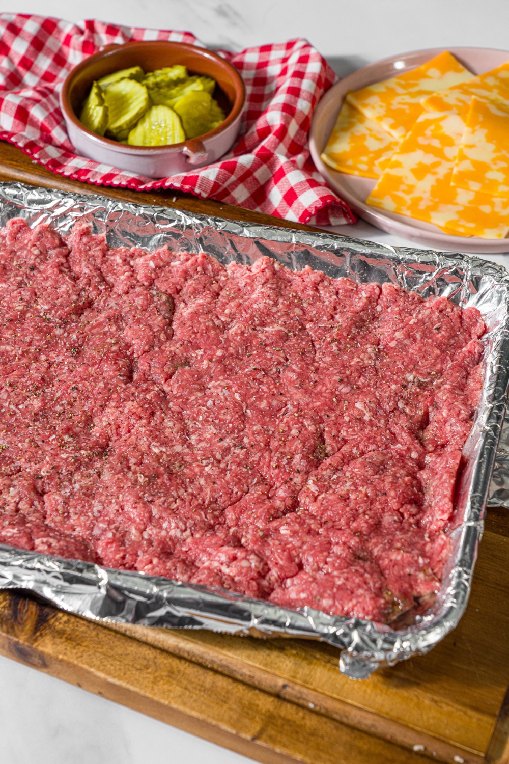 A baking sheet lined with foil with seasoned ground beef spread across the sheet. The sheet is on a wooden board.