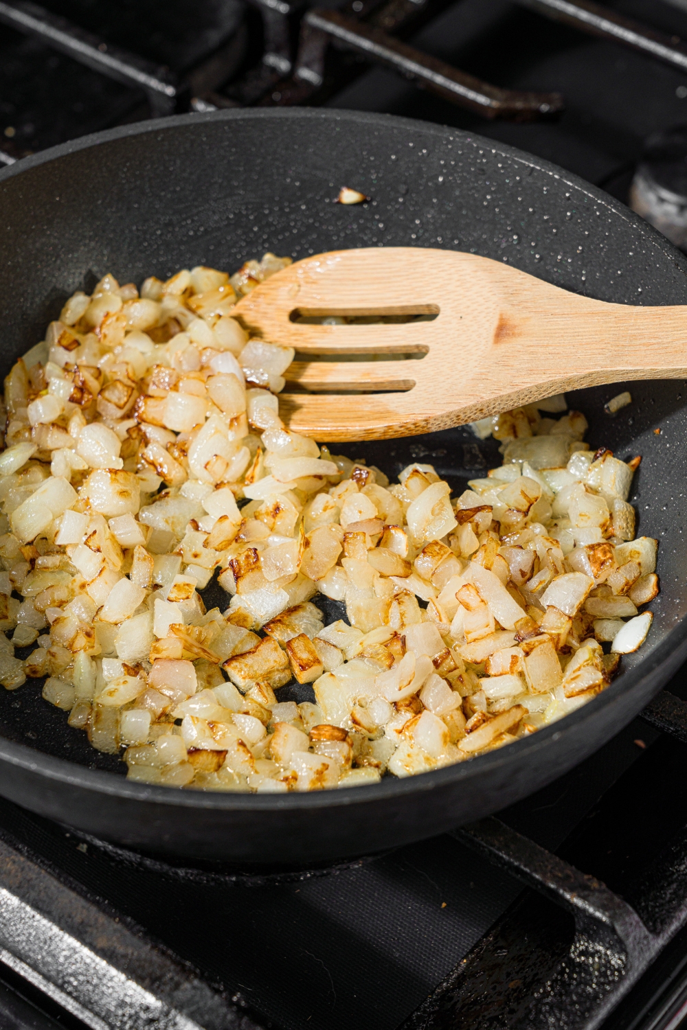 A skillet with a wooden spoon stirring sautéed diced onions. The skillet is cooking on the stove.