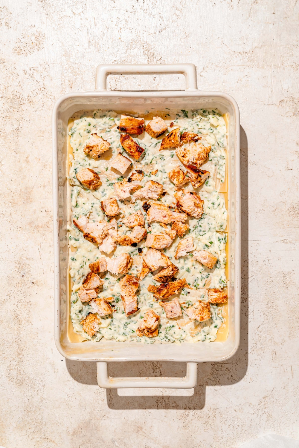 A baking dish with lasagna noodles layered with ricotta cheese mixture and cubed chicken. The dish is on a tan counter.