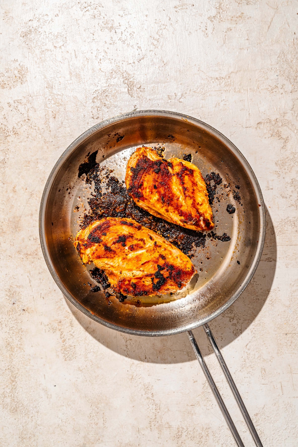 A skillet with two seared seasoned chicken breasts. The skillet is on a tan counter.