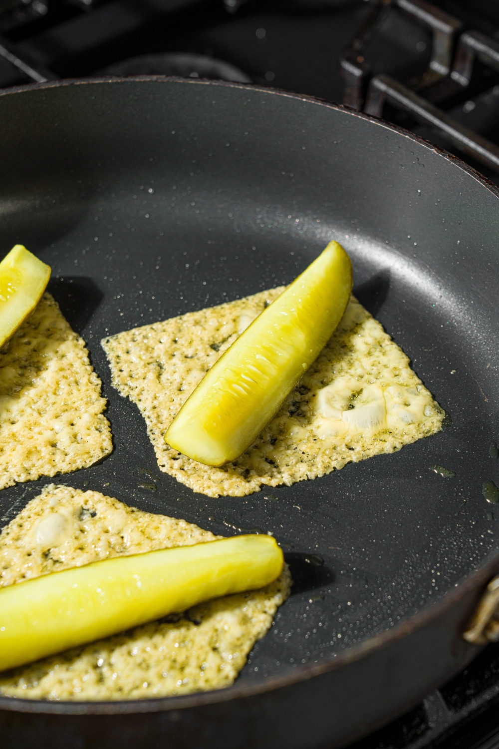 A skillet with fried slices of gouda cheese and a pickle in the center. The skillet is on a stove.