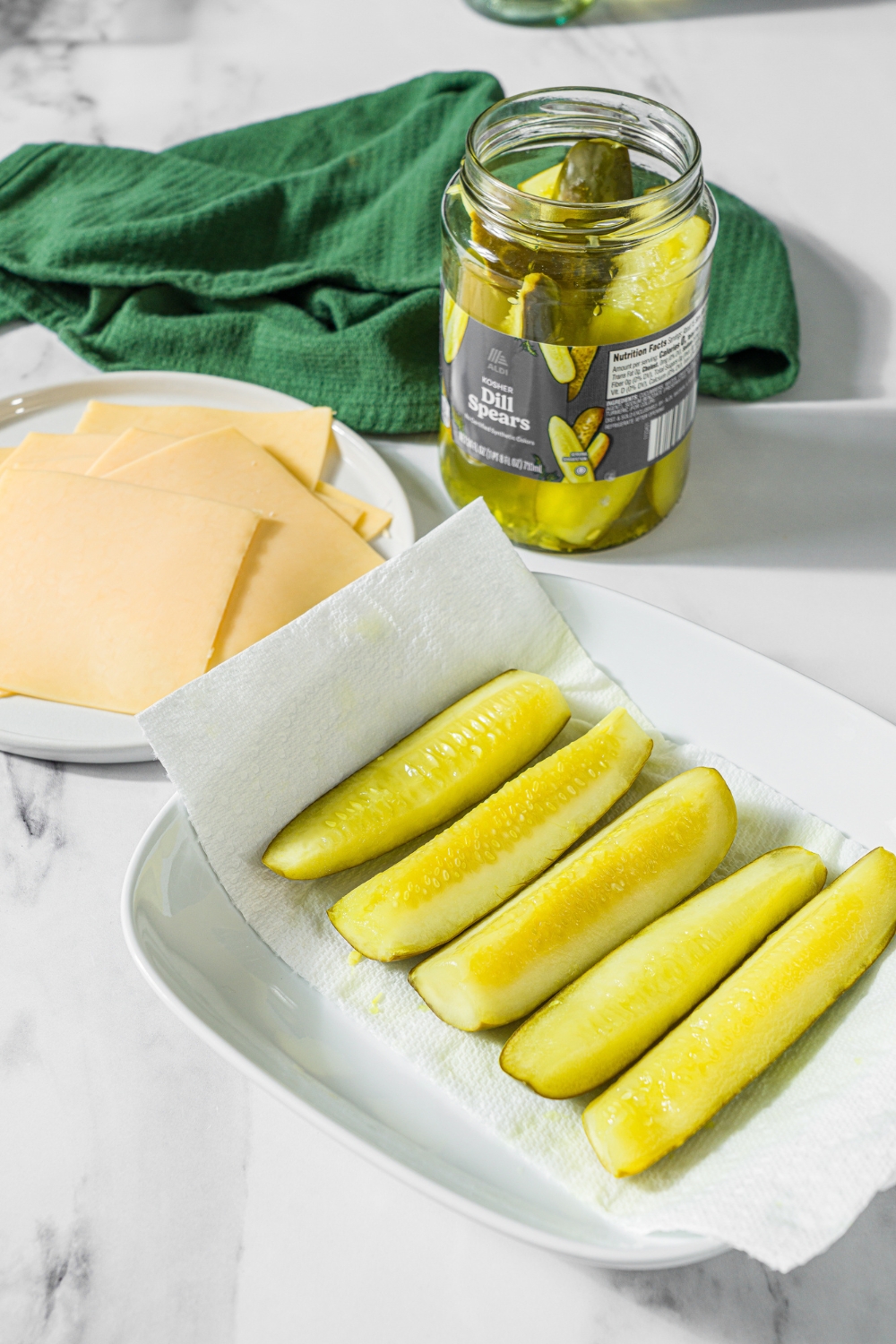 A marble counter with pickles on a white tray lined with paper towel. The tray is on a marble counter with a plate of sliced cheese and a jar of pickles.