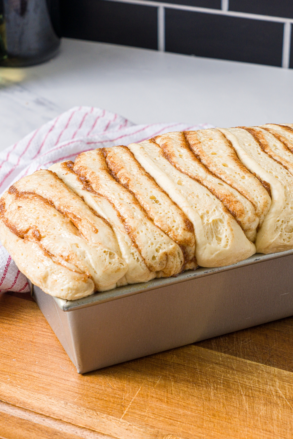 A loaf pan with risen cinnamon pull apart dough loaf. The pan is on a wooden board with a red striped napkin.