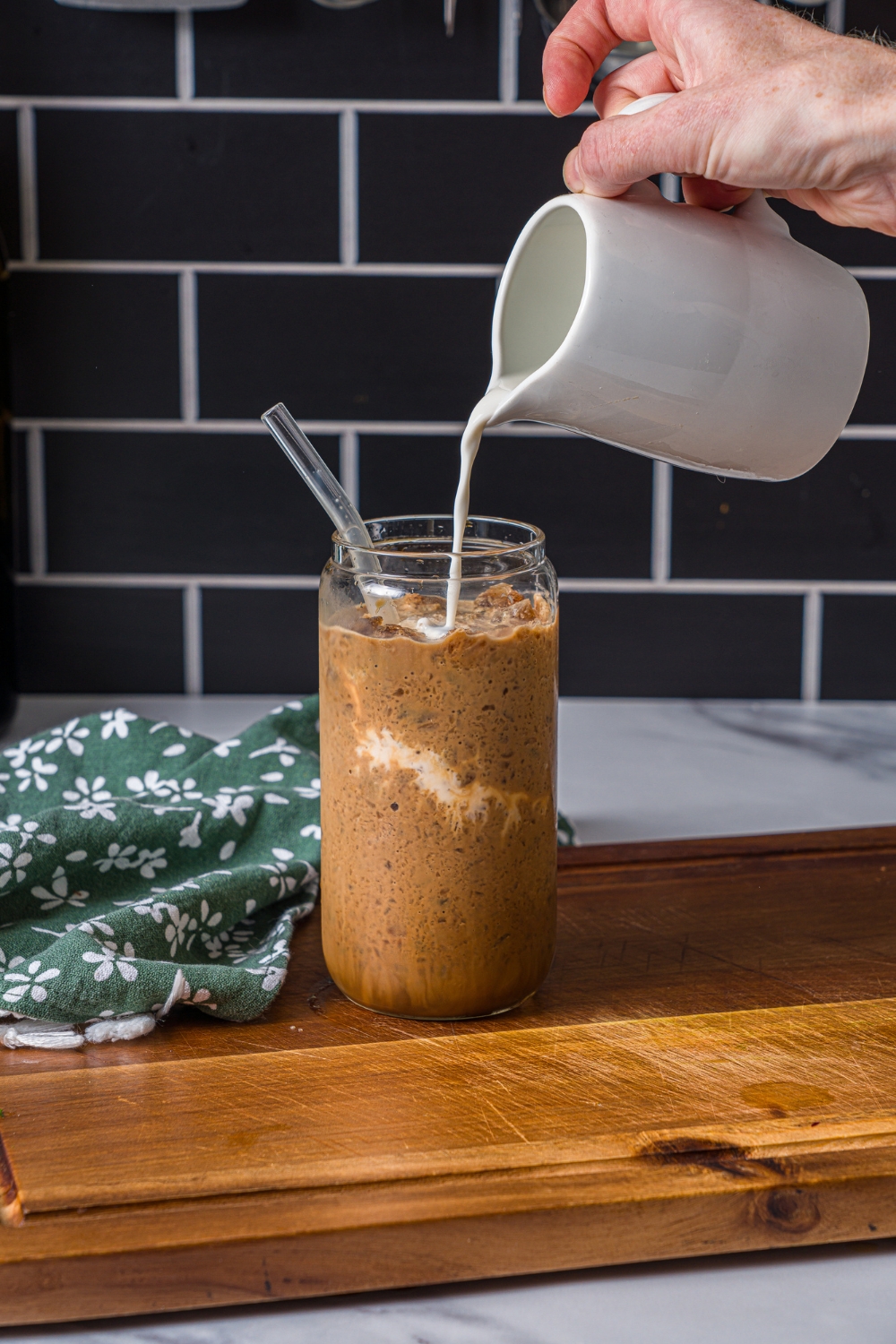 A wooden board with a glass of coffee slushie with milk being poured into the glass. The wooden board is on a marble counter with a green floral napkin.