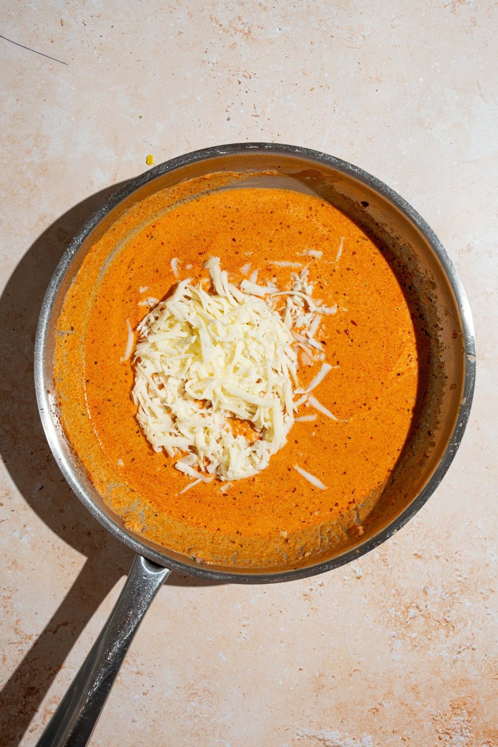 A skillet with creamy tomato sauce topped with shredded mozzarella cheese. The skillet is on a tan counter.