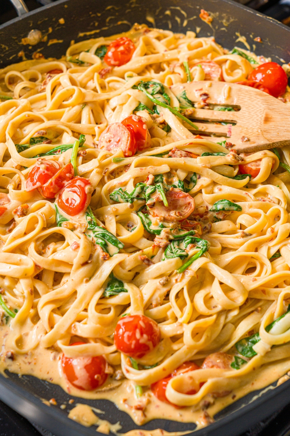 A wooden spoon stirring a creamy tuscan pasta mixture with linguine pasta, spinach, and tomatoes. The skillet is cooking on the stove.