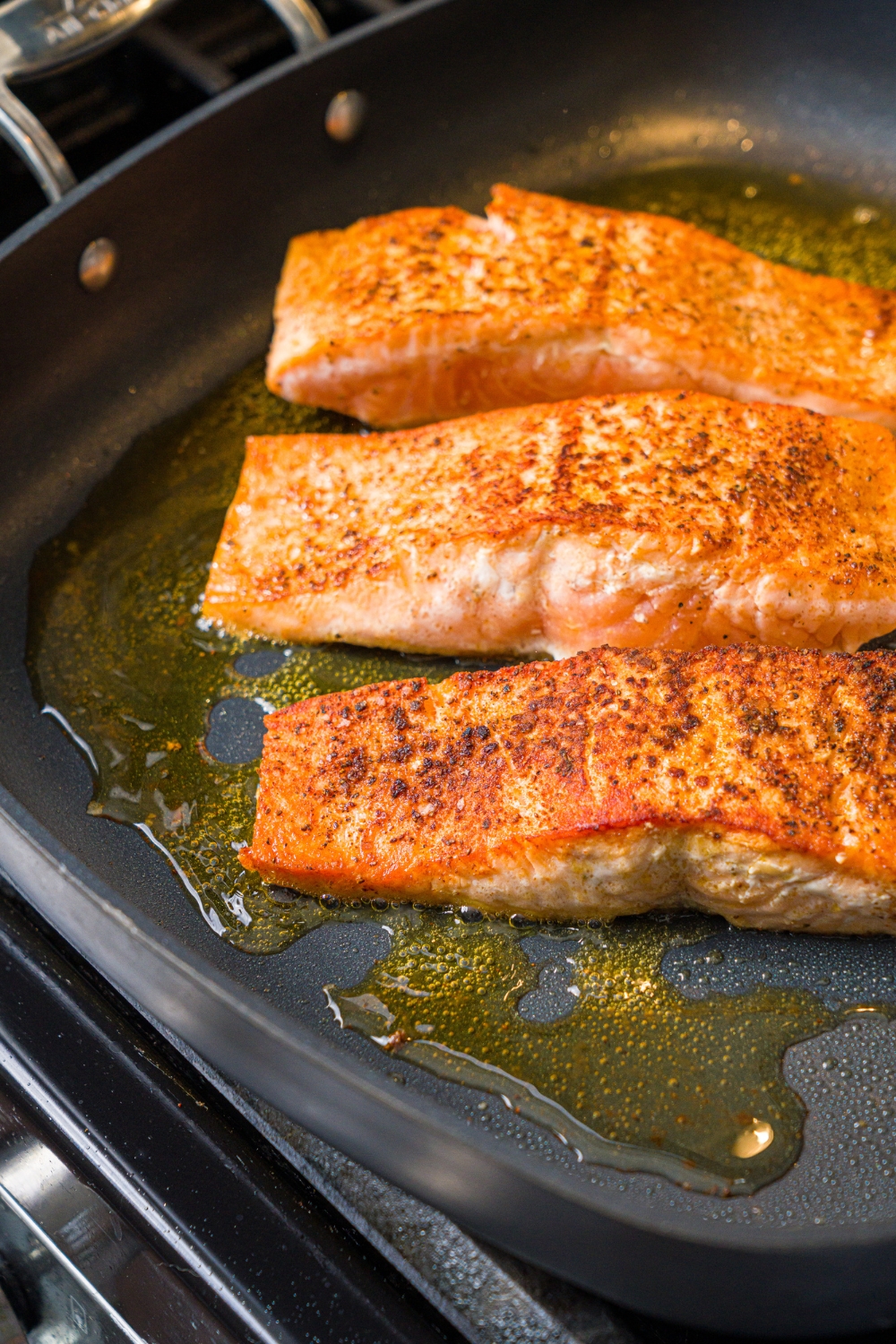 A skillet with three seared seasoned salmon with oil. The skillet is cooking on the stove.