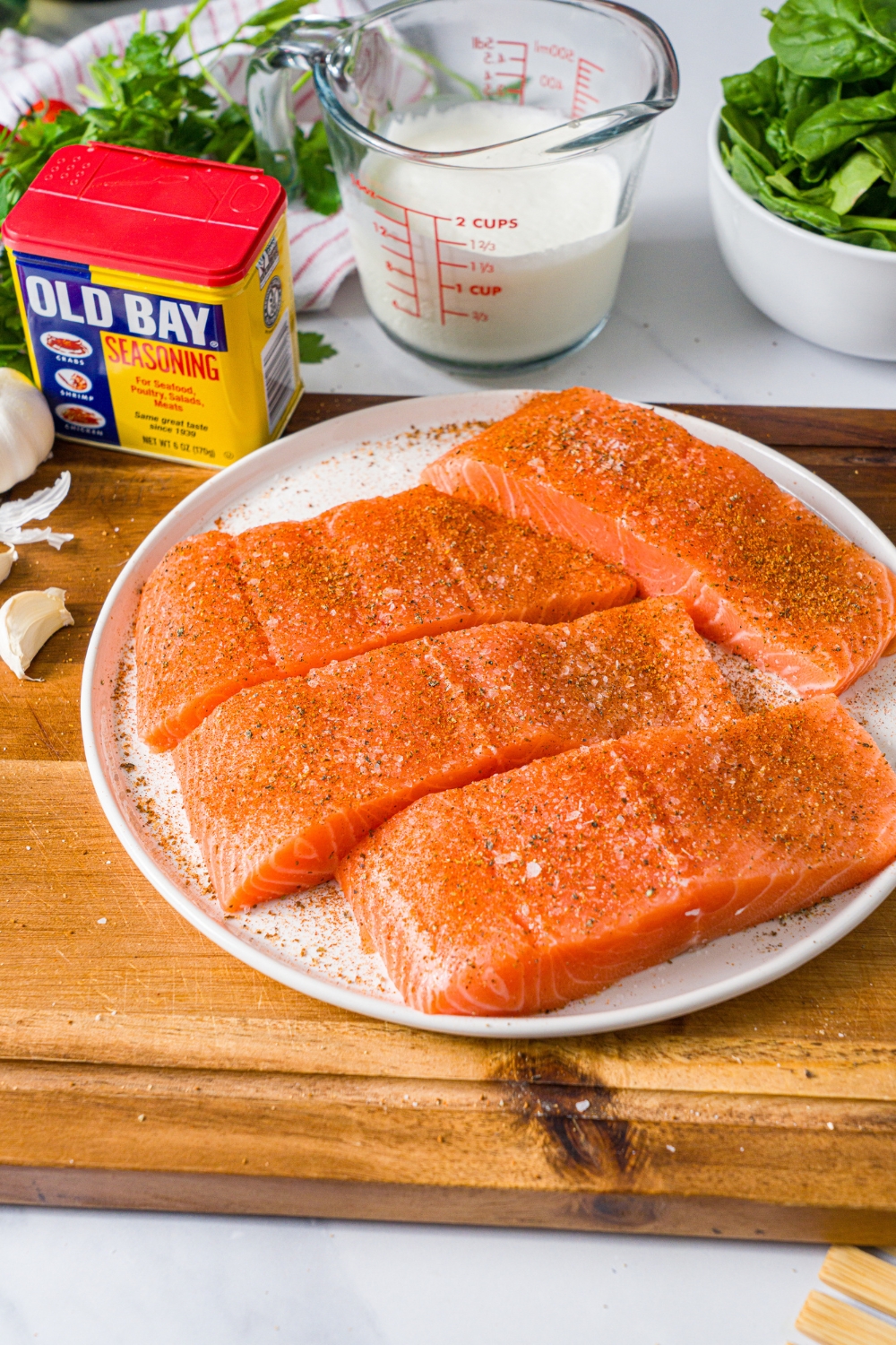 A white plate with three seasoned salmon fillets. The plate is on a wooden board on a marble counter with ingredients.