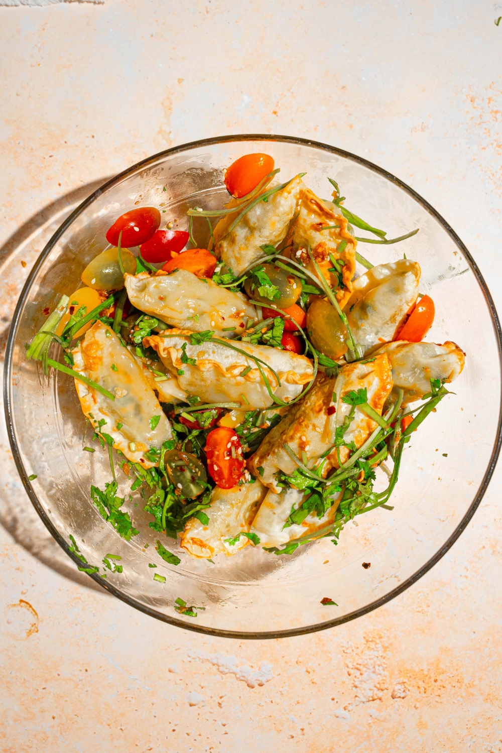 A glass bowl with crispy dumplings tossed in scallions, tomatoes, and cilantro with soy sauce and chili oil dressing. The bowl is on a tan counter.