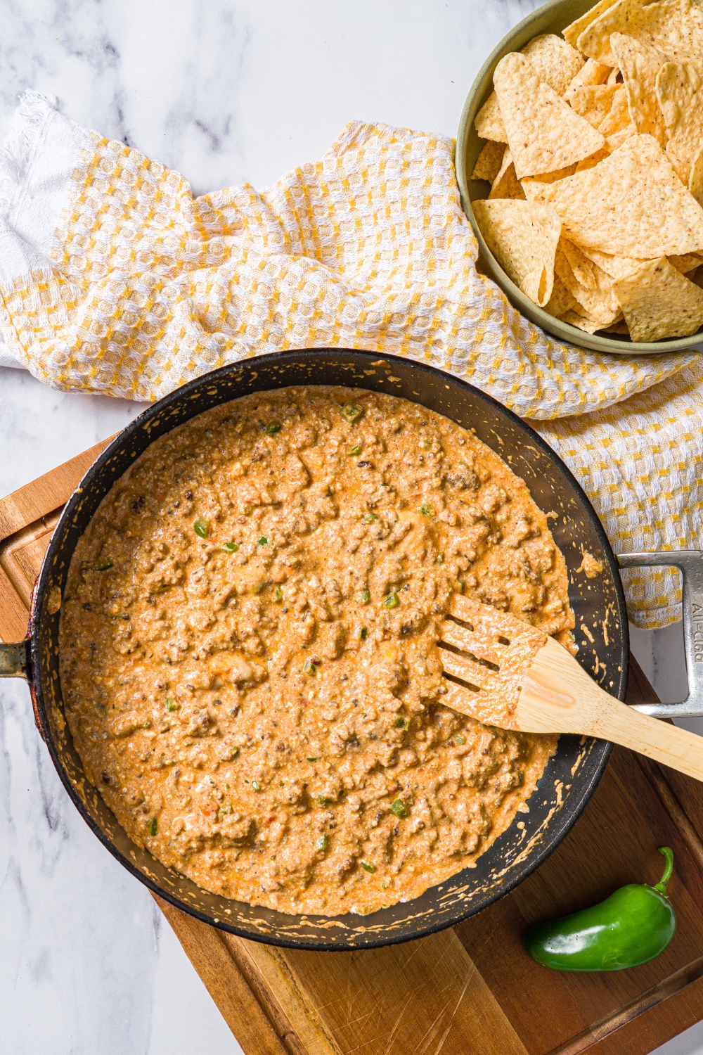 A skillet with a slotted spoon stirring divorced dad dip mixture. The skillet is on a wooden board on a marble counter with a yellow checkered napkin and a bowl of chips.