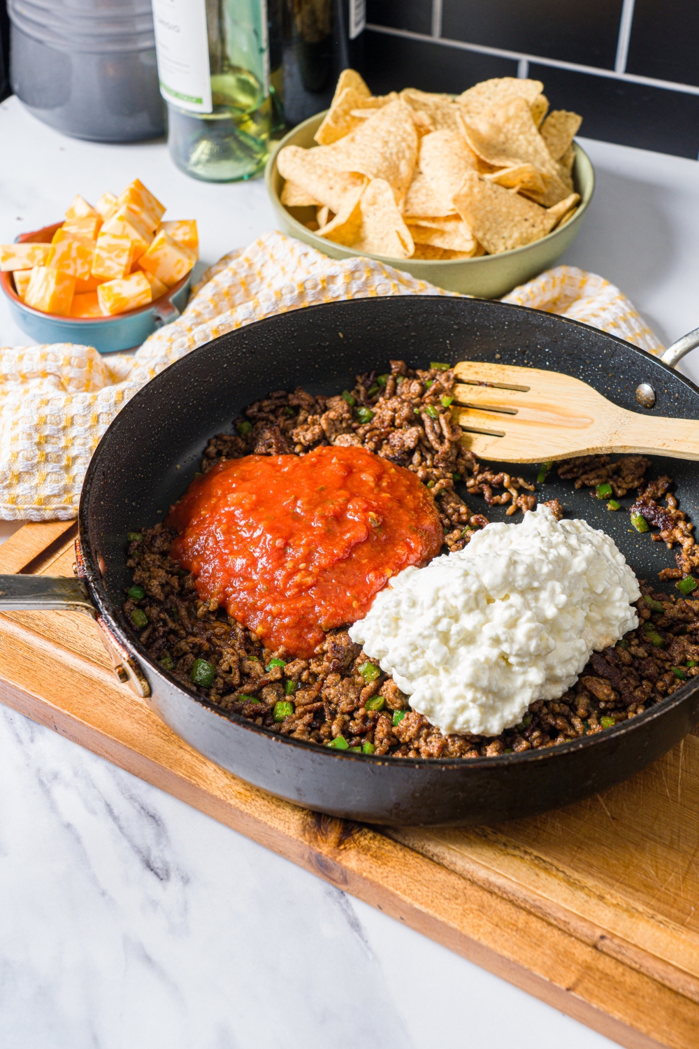 A skillet with seasoned ground beef, salsa, and cottage cheese. The skillet is on a wooden board on a marble counter with a yellow checkered napkin and bowl of chips.