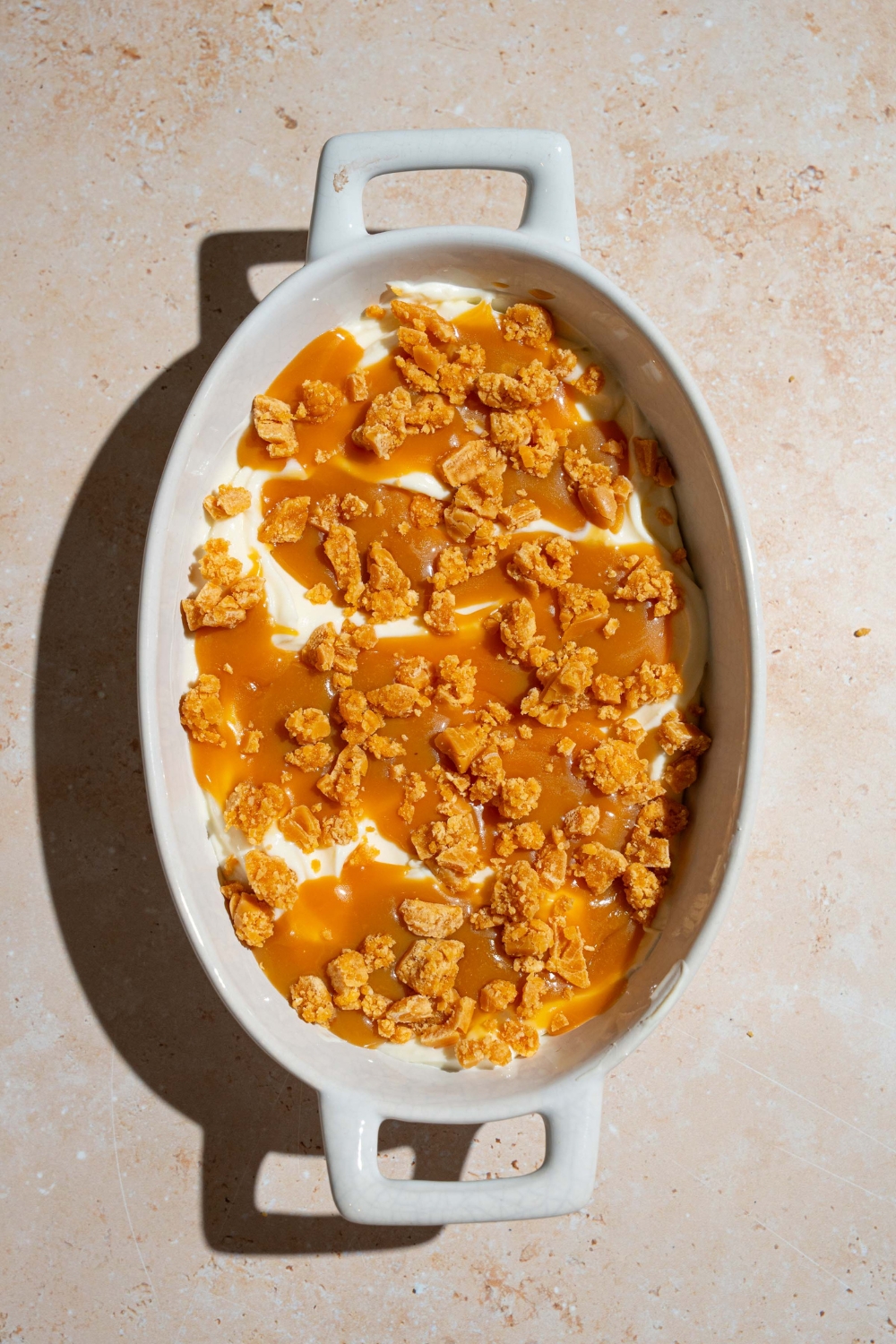 A platter covered with cheesecake filling and layered with caramel sauce and toffee bits. The platter is on a tan counter.