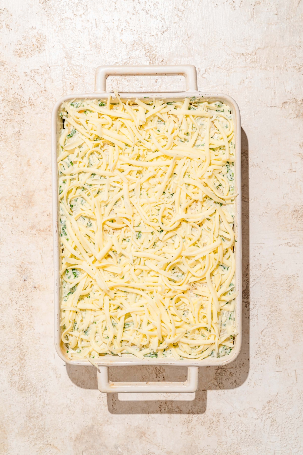 A baking dish with lasagna noodles layered with ricotta cheese mixture and cubed chicken, additional noodles, and topped with shredded mozzarella cheese. The dish is on a tan counter.