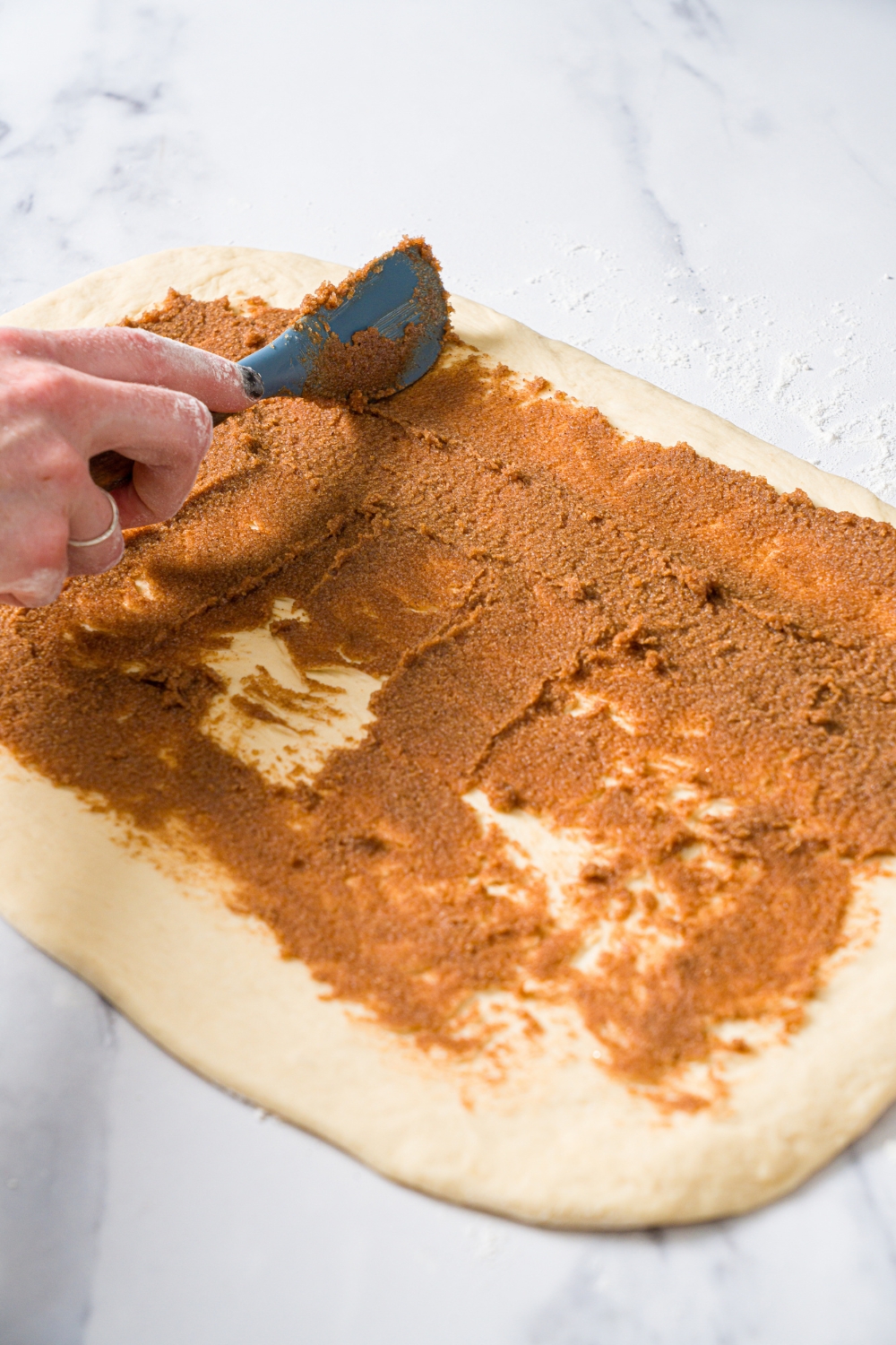 A marble counter with a cinnamon sugar mixture being spread over rolled out dough.